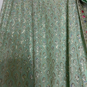 May include: A light mint green skirt featuring an intricate gold thread embroidery pattern in a repeating leaf motif. The fabric is embellished with numerous small, iridescent sequins that catch the light, and a decorative border with colorful stones.