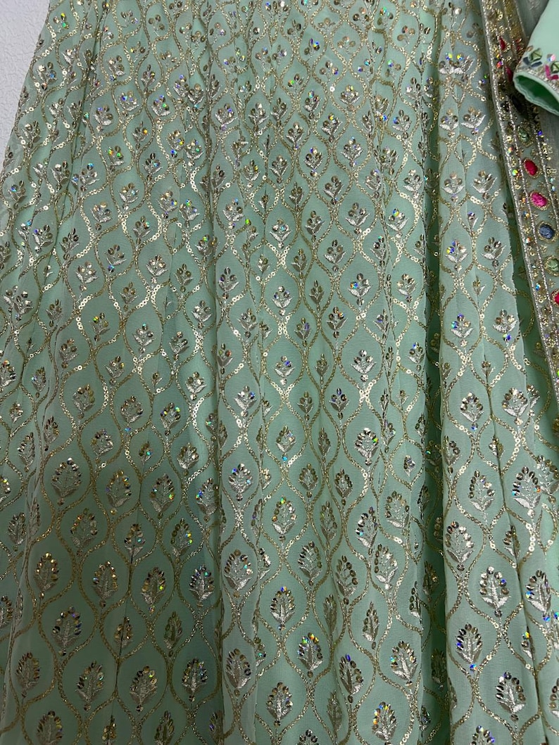 May include: A light mint green skirt featuring an intricate gold thread embroidery pattern in a repeating leaf motif. The fabric is embellished with numerous small, iridescent sequins that catch the light, and a decorative border with colorful stones.