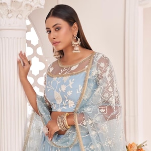May include: A light blue embroidered lehenga choli set. The top features floral embroidery and a sheer neckline. The set includes a matching dupatta with intricate detailing. Gold bangles and earrings complete the look. The number "2112" is visible.