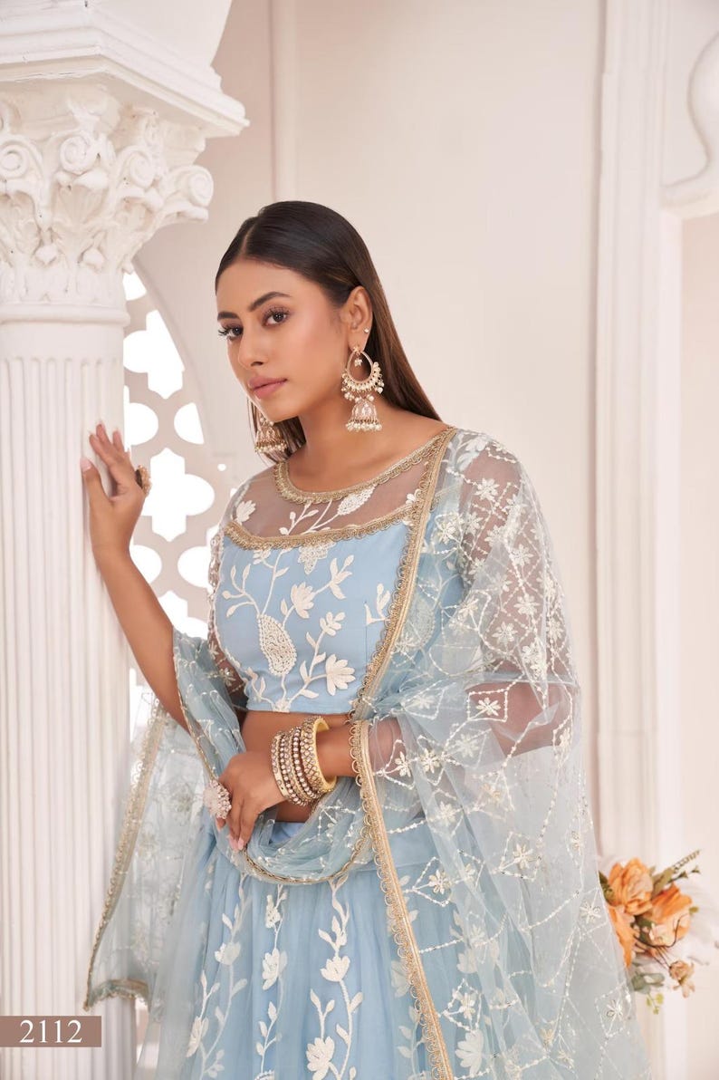 May include: A light blue embroidered lehenga choli set. The top features floral embroidery and a sheer neckline. The set includes a matching dupatta with intricate detailing. Gold bangles and earrings complete the look. The number "2112" is visible.