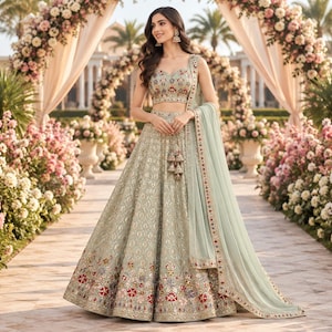 May include: A light sage green lehenga choli featuring a sleeveless blouse and a floor-length skirt with intricate embroidery. The ensemble includes a matching sheer dupatta with a decorative border, adorned with floral patterns in red, blue, and gold.