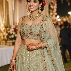 May include: A sage green lehenga set featuring a sleeveless blouse and a floor-length skirt with intricate gold embroidery and floral accents. The ensemble includes a matching sheer dupatta with a decorative border and metallic embellishments.