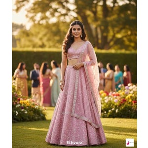 May include: A woman in a light pink lehenga, a traditional Indian outfit, poses outdoors. The outfit includes a crop top, a long skirt, and a sheer scarf. The lehenga is embellished with delicate patterns. The background features a garden and a group of people.