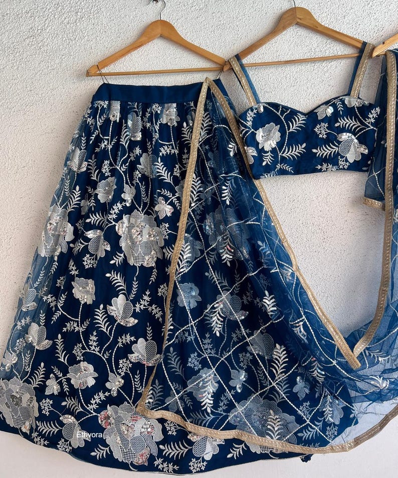 May include: A navy blue embroidered lehenga set with silver floral detailing. The set includes a skirt, a fitted top, and a sheer dupatta with gold trim. The outfit is displayed on wooden hangers against a white background.
