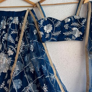 May include: A navy blue embroidered ensemble featuring a crop top, skirt, and sheer scarf. The set is adorned with silver floral embroidery and gold trim. The outfit is displayed on wooden hangers against a neutral background.