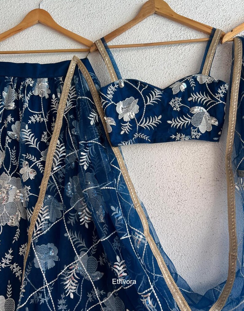 May include: A navy blue embroidered ensemble featuring a crop top, skirt, and sheer scarf. The set is adorned with silver floral embroidery and gold trim. The outfit is displayed on wooden hangers against a neutral background.
