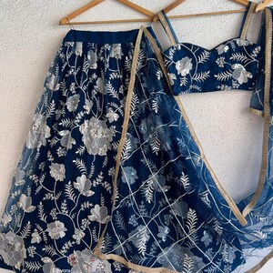 May include: A navy blue embroidered lehenga set with silver floral detailing. The set includes a skirt, a fitted top, and a sheer dupatta with gold trim. The outfit is displayed on wooden hangers against a white background.