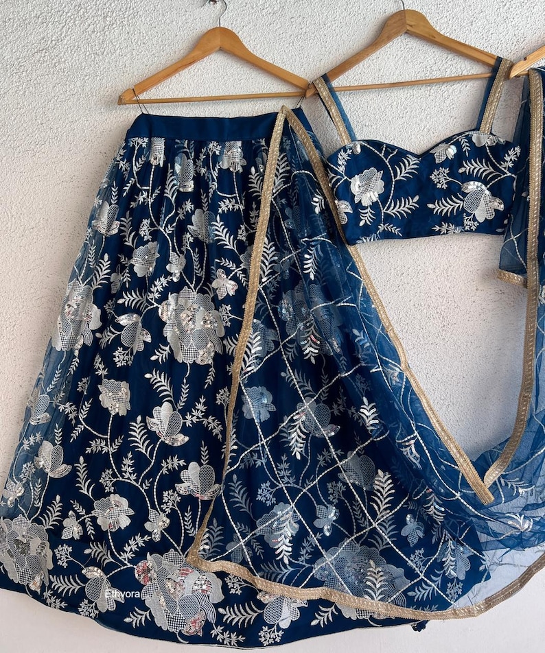 May include: A navy blue embroidered lehenga set with silver floral detailing. The set includes a skirt, a fitted top, and a sheer dupatta with gold trim. The outfit is displayed on wooden hangers against a white background.