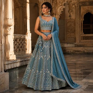 May include: A dusty blue lehenga choli set featuring a sleeveless blouse and a floor-length skirt, both heavily embellished with intricate silver embroidery and mirror work. A matching sheer dupatta with a silver border is draped over one shoulder.
