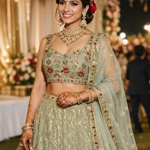 May include: A sage green lehenga set featuring a sleeveless blouse and a floor-length skirt with intricate gold embroidery and floral accents. The ensemble includes a matching sheer dupatta with a decorative border and metallic embellishments.