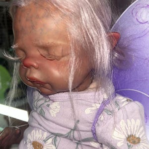 May include: A lifelike baby doll with pale skin, closed eyes, and long eyelashes. The doll has light purple hair and is wearing a lavender onesie with a daisy floral pattern. Purple fairy wings are visible.