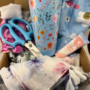 May include: A gift box with a light blue floral tumbler, blue scissors, hand cream, a floral scarf with blue tassels, and a floral patterned hat. The items are in a cardboard box.