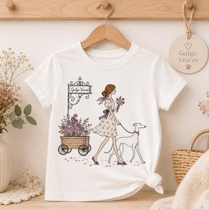 以下が含まれることがあります： 白いTシャツに、気まぐれなイラストが描かれています。花柄のドレスを着た女性が、花を持ちながらグレイハウンドと散歩しています。「Galgo Voices」と書かれた看板と紫の花でいっぱいの木製のカートも描かれています。