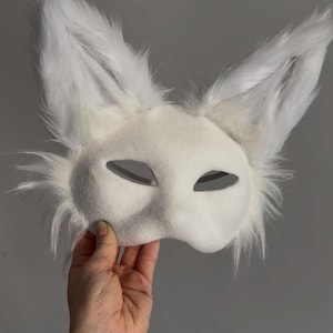 May include: A white fox mask with large, upright ears and a fluffy, textured appearance. The mask has eye cutouts and is held up against a gray background. The mask is likely for costume or cosplay purposes.