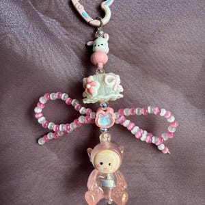 May include: A pink and white beaded keyring with a heart-shaped clasp. The keyring features a small bunny, a cake, a bow, and a translucent pink figure. The background is a purple fabric.