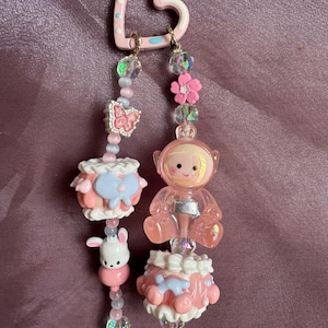 May include: Pink and blue beaded earrings with a heart-shaped top. One earring features a pink astronaut charm, while the other has a white bunny charm. Both earrings have pink, blue, and clear beads and decorative charms.