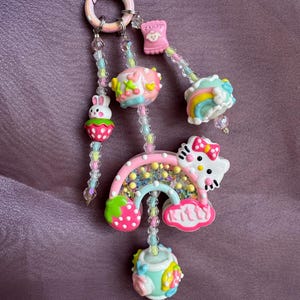 Puede incluir: Un llavero colorido con varios dijes. Los dijes incluyen un arcoíris, una fresa, un conejito, un envoltorio de caramelos y un diseño de Hello Kitty. El llavero está adornado con cuentas iridiscentes y un anillo rosa.