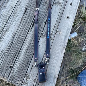 May include: A dark brown leather radio strap with red stitching and silver-tone hardware. The strap has the text "GRAPEDUDE" in blue and yellow. A small black leather case is attached to the strap.