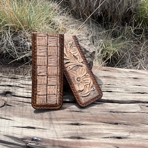 May include: Two brown leather cases. One has a woven pattern, the other a floral design. Both have a dark brown border. The cases are displayed on a weathered wooden surface, with dry grass and rocks in the background.