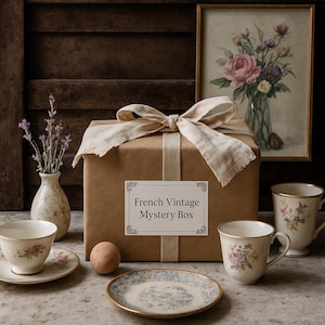 Könnte beinhalten: Eine braune, mit Papier umwickelte Schachtel, die mit einem beigen Band verschnürt ist und mit "French Vintage Mystery Box" beschriftet ist. Die Szene umfasst eine Teetasse, eine Untertasse und eine kleine Vase mit Lavendelzweigen. Ein gerahmter Blumendruck und zwei Teetassen sind ebenfalls zu sehen.
