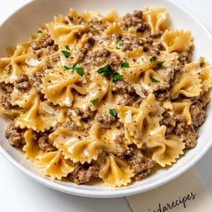 May include: A white bowl filled with bow tie pasta coated in a creamy brown meat sauce, topped with flecks of black pepper and green herbs. A small piece of paper with the text "jadarecipes" written in black cursive script is visible below the bowl.