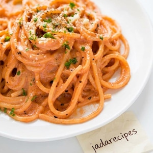 Puede incluir: Un plato de espaguetis cubiertos con una salsa de tomate cremosa de color naranja, espolvoreados con queso rallado, pimienta negra y perejil verde fresco. Una pequeña nota cuadrada con el texto manuscrito jadarecipes está junto al plato.