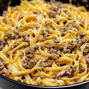 May include: Close-up of a pan filled with creamy, golden-yellow fettuccine pasta, mixed with ground meat. The pasta is garnished with small green herbs, creating a delicious and appetizing meal.
