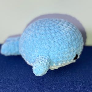 May include: A light blue crocheted whale plush toy with a white belly and small black eyes. The whale is lying on its side on a blue surface. The plush toy is made of soft, textured yarn.