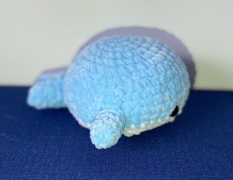 May include: A light blue crocheted whale plush toy with a white belly and small black eyes. The whale is lying on its side on a blue surface. The plush toy is made of soft, textured yarn.