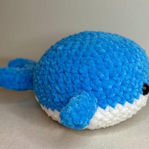 May include: A plush, blue whale toy with a white belly. The whale has small, black eyes and a textured, fuzzy surface. The flippers and tail are also blue. This handmade toy is suitable for children.