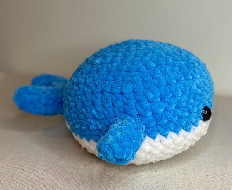 May include: A plush, blue whale toy with a white belly. The whale has small, black eyes and a textured, fuzzy surface. The flippers and tail are also blue. This handmade toy is suitable for children.