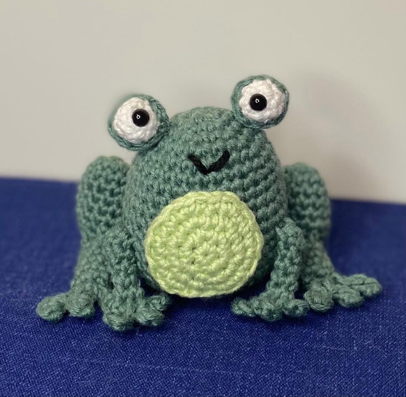 Op de afbeelding: Een handgemaakte gehaakte kikker in groen- en lichtgroentinten. De kikker heeft grote, witgerande ogen met zwarte pupillen, een lichtgroene buik en gedetailleerde poten. De kikker zit op een blauw oppervlak.