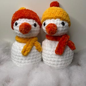 May include: Two handmade crochet snowmen. Each snowman has a white body, orange carrot nose, and black eyes. One wears an orange hat and yellow scarf, the other a yellow hat and orange scarf. They are on a white surface.