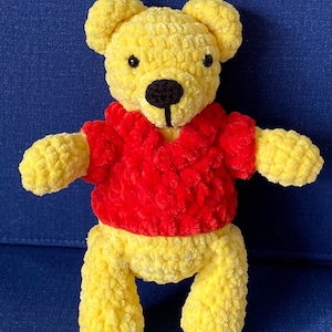 May include: A handmade yellow crochet bear wearing a red shirt. The bear has black eyes and a black nose. The plush toy is standing on a blue surface. The bear's arms and legs are extended.