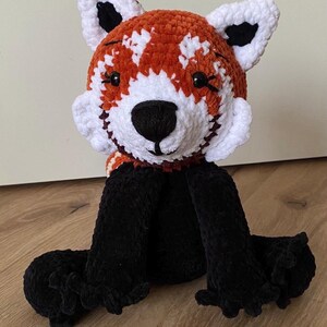 May include: A handmade crochet red panda plush toy. The plush features a head with white and orange fur, black eyes, and a black nose. The ears are white with black tips, and the body and legs are black. The toy is sitting on a wooden surface.