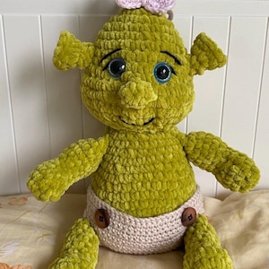 May include: A green crocheted plush doll with a textured yarn finish, featuring large blue eyes, prominent ears, and a small nose. The doll wears a light beige crocheted diaper secured with two brown buttons and has a small pink bow on its head.