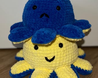 Op de afbeelding: Een handgemaakte, omkeerbare gehaakte octopus knuffel. De ene kant is geel met een lachend gezicht en blauwe accenten, de andere is blauw met een verdrietig gezicht. De knuffel is zacht en knuffelig, perfect voor kinderen of als decoratie.