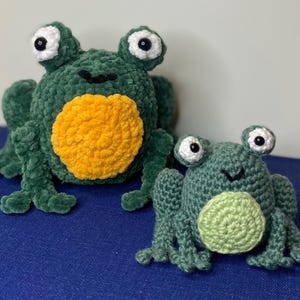 May include: Two handmade crochet frogs in shades of green, with large white and black eyes. One frog is larger, with a yellow belly, and the other is smaller, with a light green belly. The frogs are sitting on a blue surface.