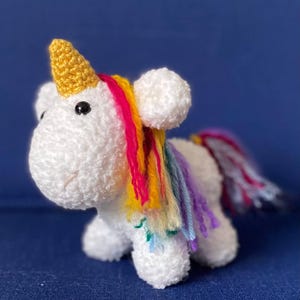 May include: A white crocheted unicorn toy with a golden horn, black eyes, and a pink stitched mouth. The unicorn has a rainbow mane and tail. The toy is made of soft, textured yarn and sits on a blue surface.