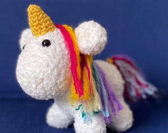 Op de afbeelding: Een wit gehaakt eenhoornspeelgoed met een gouden hoorn, zwarte ogen en een roze gestikte mond. De eenhoorn heeft een regenboogmanen en -staart. Het speelgoed is gemaakt van zacht, getextureerd garen en ligt op een blauw oppervlak.