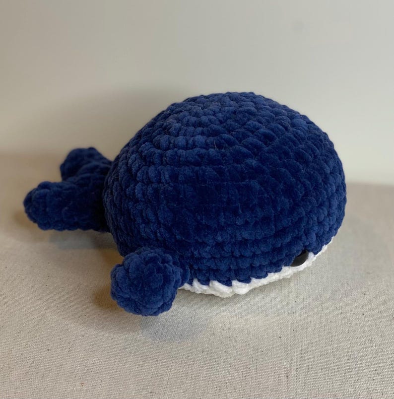 May include: A plush, navy blue whale toy with a rounded body and small fins. The whale is made of a soft, textured material. The underside is white with black accents. A handmade, cuddly ocean-themed toy.