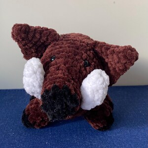 May include: A handmade, plush, brown crocheted boar with white tusks and black accents. The boar has small black eyes and large ears. The plush toy is sitting on a blue surface.