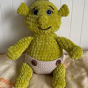 May include: A handmade crochet plush doll featuring a bright green textured yarn body, large black plastic eyes, and a small tuft of light brown yarn on top of its head. The doll wears a beige crocheted diaper secured with two dark brown buttons.
