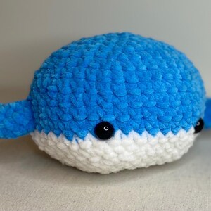 May include: A handmade, plush whale toy in shades of blue and white. The whale features a rounded body, small fins, and black button eyes. The toy is made of soft, textured yarn, giving it a cuddly appearance. A charming, handcrafted item.