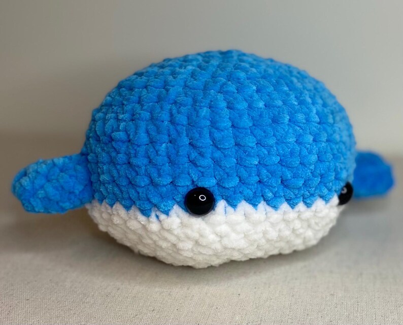 May include: A handmade, plush whale toy in shades of blue and white. The whale features a rounded body, small fins, and black button eyes. The toy is made of soft, textured yarn, giving it a cuddly appearance. A charming, handcrafted item.