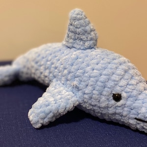 May include: A light blue crocheted dolphin plush toy with a dorsal fin, flippers, and a tail. The dolphin has a black eye and a stitched mouth. The plush is lying on a dark blue surface.