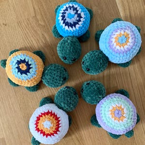 May include: A collection of six handmade crochet turtles. Each turtle features a green body and flippers, with a round, colorful shell. Shell colors include blue, yellow, white, and purple, with intricate patterns. The turtles are arranged on a wooden surface.