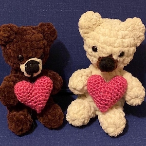 May include: Two crocheted teddy bears, one brown and one cream, each holding a pink heart. The bears have black eyes and noses, and are made of a soft, textured yarn. The background is a solid blue.