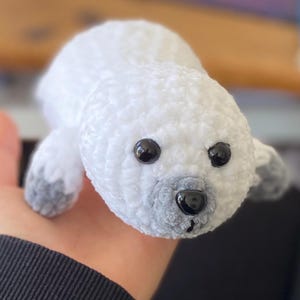 May include: A small, white, crocheted plush seal toy with a rounded body and head. It features two shiny black plastic eyes and a small black plastic nose set against a light gray muzzle. The flippers are tipped with a soft, light gray yarn.