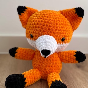 May include: A handmade orange and white crocheted fox plush toy with black accents on the ears, paws, and nose. The fox has a white muzzle and black eyes. The plush is sitting on a wooden surface.